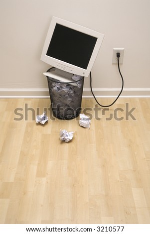 Computer monitor in trash can surrounded by crumpled up paper.