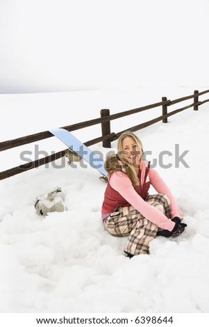 Young woman in winter clothes by fence in snowy field kneeling by snowboard and smiling.