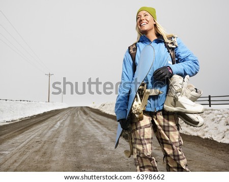 Young woman in winter clothes standing on muddy dirt road holding snowboard and boots smiling.