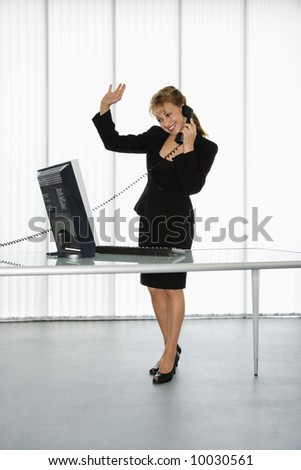 Caucasian businesswoman standing at computer desk waving on telephone.