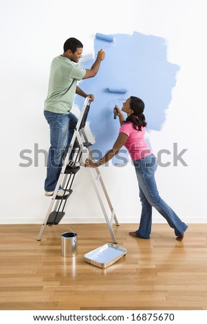 African American male and female couple painting wall blue and smiling at each other.