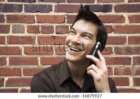 Smiling young Asian man next to brick wall talking on cell phone.