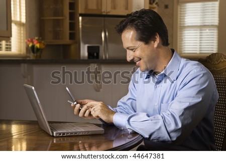 Man sitting at table smiling with cell phone and laptop. Horizontally framed shot.