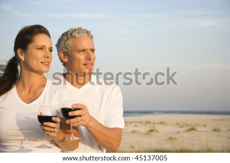 Couple drink red wine at the beach and gaze into the distance together. Horizontal shot.