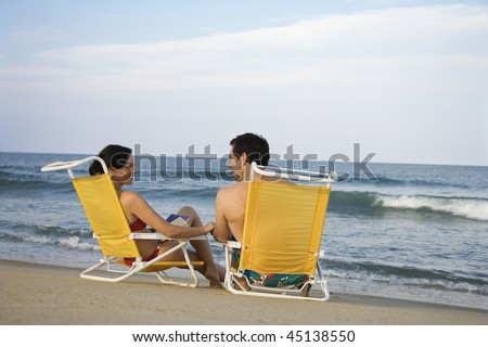 Smiling couple sit in chairs on the beach as the waves come in. Horizontal shot.