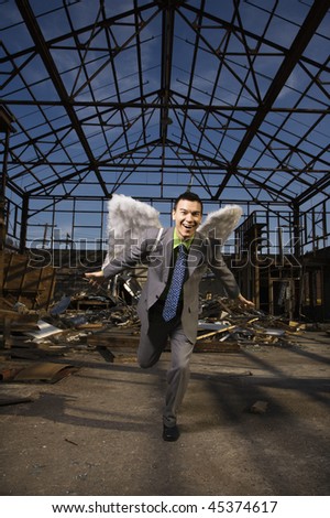 Young businessman smiles with angel wings on his back and attempts takeoff in an abandoned building. Vertical shot.
