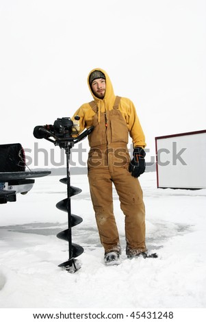 Young man in snow gear poses with an ice drill in a winter environment. Vertical shot.