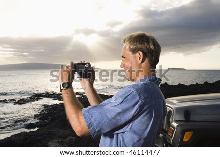 Side view of a smiling Caucasian man photographing a scenic sunset at a beach. Horizontal format.