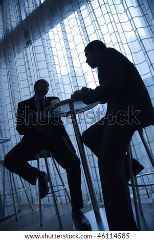 Tilt view silhouette of African-American and Asian businessmen sitting at a table and having coffee in front of a curtained window. Vertical format.