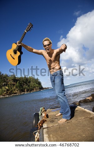 Shirtless young man wearing sunglasses, standing on a pier and holding a guitar up in the air with an enthusiastic expression. Vertical shot.
