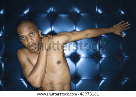 Handsome young man sitting with his hand on his shoulder against a blue padded background. He is shirtless and has one arm outstreatched. Horizontal shot.