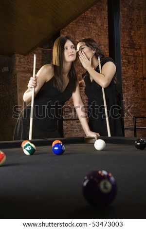 Two attractive young women at pool table as one whispers into other\'s ear.