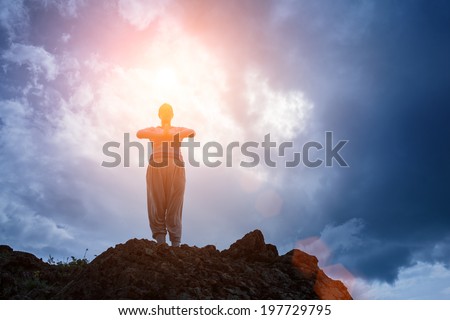 Woman standing on top of mountain doing yoga meditation. Stormy sky background, sun shining on her.