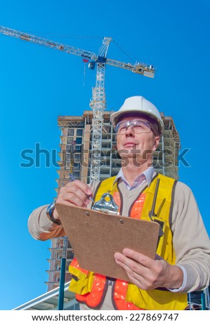 Engineer, construction worker making notices, calculations with high-rise building under construction as a background. The site with cranes against blue sky. Vancouver, Canada. Vertical.
