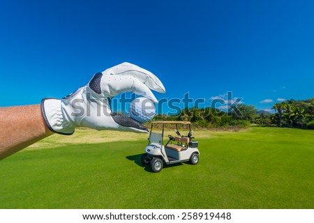 Hand wearing golf glove, holding golf ball over beautiful golf course with the golf cart at the back.