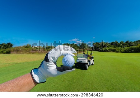 Hand wearing golf glove holding golf ball over beautiful course with blue sky. Vertical.