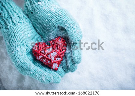 Woman hands in light teal knitted mittens are holding a beautiful glossy red heart in a snow background. Love and St. Valentine concept.