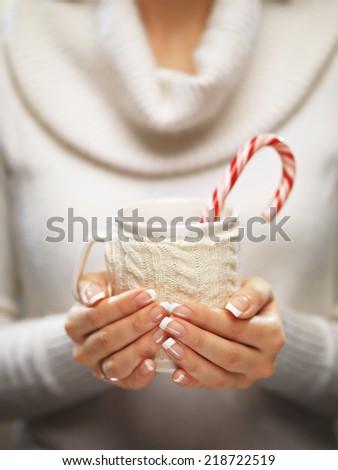 Woman holds a winter cup close up. Woman hands with elegant french manicure nails design holding a cozy knitted mugwith cocoa, tea or coffee and a candy cane. Winter and Christmas time concept.