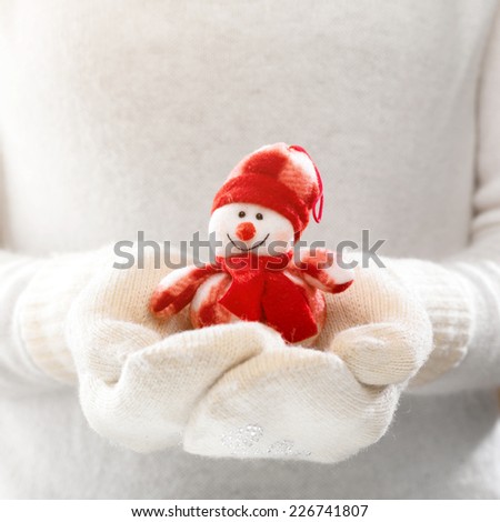 Female hands holding a cute happy Snowman. Woman hands in teal mittens showing a Snowman gift dresses in red hat and scarf. Cute Christmas present. Winter holidays concept.