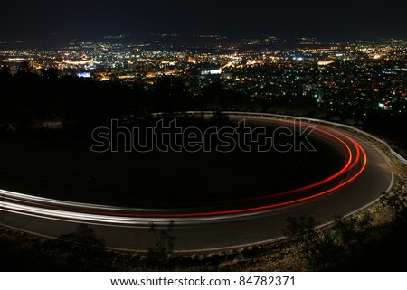Nighttime, long exposure of car lights around hairpin curve on mountain road with city lights gleaming below.