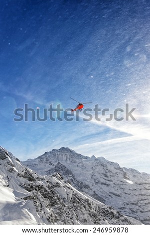 Red helicopter in flight in winter swiss alps with snow powder streams