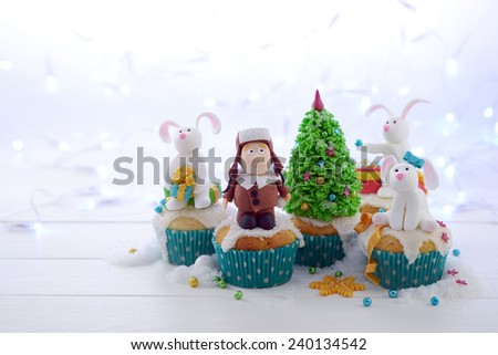 Festive cupcakes with sugar figures on a bright lights background.  A kid in a fur hat and rabbits decorating the Christmas tree.