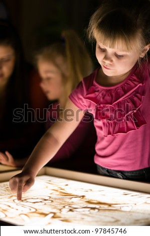 Girl paint with sand on table by her finger in classroom with friends