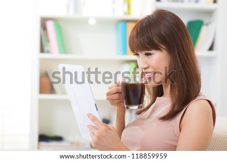 Asian female enjoying cup of coffee and digital tablet inside house