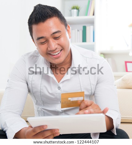 Southeast Asian man online shopping, hands holding credit card and computer tab sitting at home.