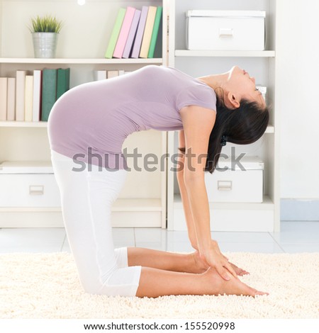 Maternity health concept. Full length healthy 8 months pregnant calm Asian woman meditating or doing yoga exercise at home. Relaxation. Yoga camel pose.