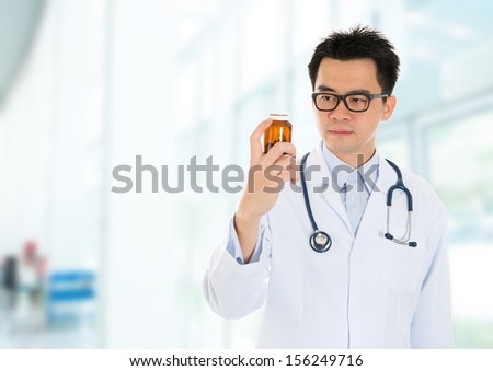 Asian male medical doctor holding a bottle of drugs, standing inside hospital building.