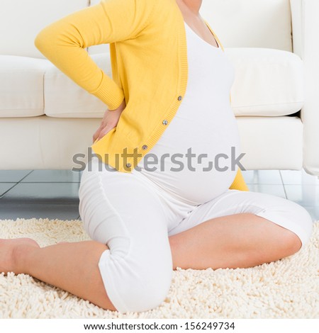 Back pain. 8 months pregnant woman holding her back while sitting on a floor at home.