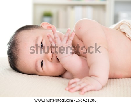 Adorable six months old Asian baby girl lying on bed biting fingers.