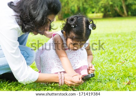 Cute Indian girl peeking through magnifying glass with parent on green lawn. Mother and daughter exploring nature at outdoor garden.