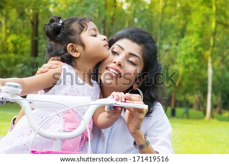 Indian family outdoor activity. Asian Mother teaching daughter cycling at the park in the morning. Child kissing parent.