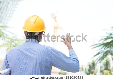 Rear view of Indian male site contractor engineer with hard hat pointing to a crane at construction site.
