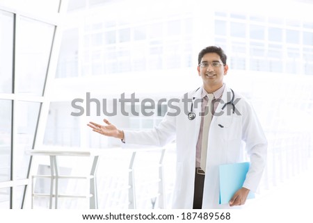 Portrait of a smiling Asian Indian male medical doctor standing inside hospital, holding file folder and showing welcome hand sign.