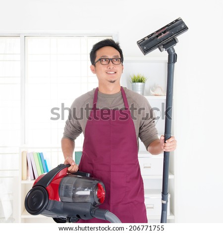 Asian man holding vacuum. House husband doing house chores, house interior.