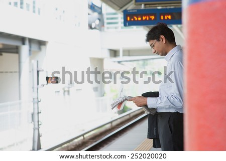 Candid Asian Indian businessman waiting at public train station, standing and reading on newspaper.