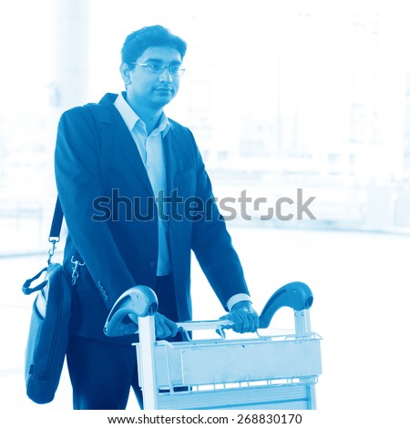 Asian Indian businessman walking with airport trolley, in blue tone.