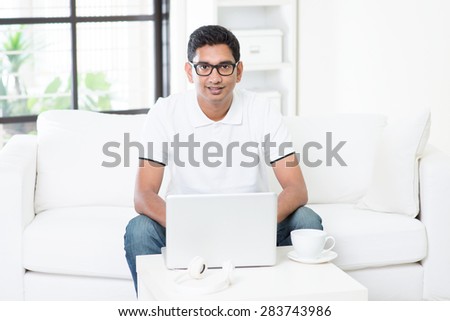 Young Indian guy start his new online business with laptop computer from home. Asian man using internet indoor, relaxed and sitting on sofa. Handsome male model working indoors.