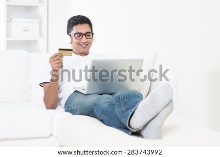 Indian guy making online credit card payment with laptop computer at home. Asian man internet shopping indoor  relaxed and sitting on sofa. Handsome male model.