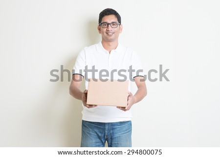 Courier delivery service concept. Indian man received a brown box, standing on plain background with shadow. Asian handsome guy model.