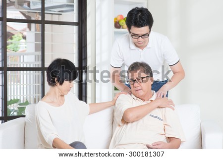 Asian old man shoulder pain, sitting on sofa with wife, son massaging father shoulder. Chinese family, senior retiree indoors living lifestyle at home.