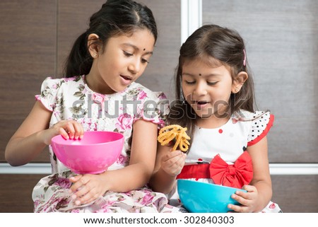 Two cute Indian girls eating traditional snack murukku. Asian sibling or children enjoying food, living lifestyle at home.