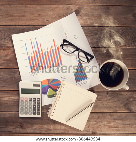Top view workspace with booklet, pen, glasses, cup of coffee and graphs. Wooden table background in vintage toned.