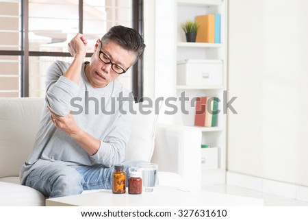 Portrait of casual 50s mature Asian man elbow pain, pressing on elbow joint with painful expression, sitting on sofa at home, medicines and water on table.