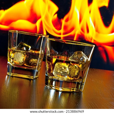 two glasses of whiskey with ice cubes in front of the flame on wood table