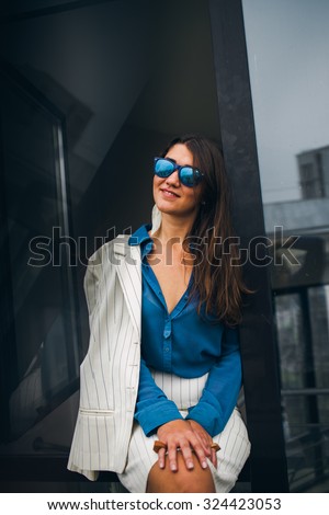 Close portrait of a young woman, she is smiling. Blue blouse, white skirt. Glasses, sunny day, office.