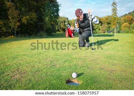 Female - golf player on knees and arms raised with putter in hand in winner pose on golf green being overjoyed as golf ball drops into hole